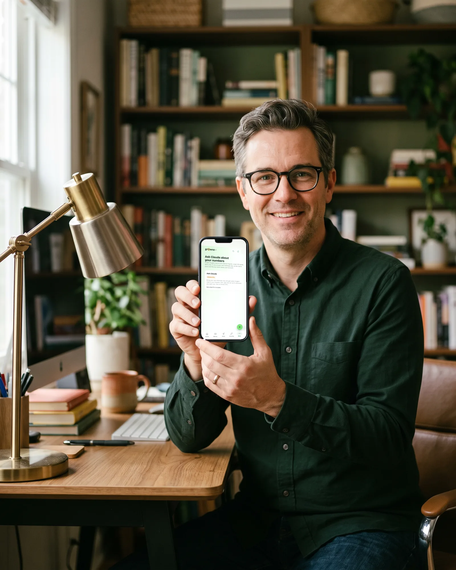 A man with glasses at a home desk holding his phone up to the camera, the assistant screen showing
